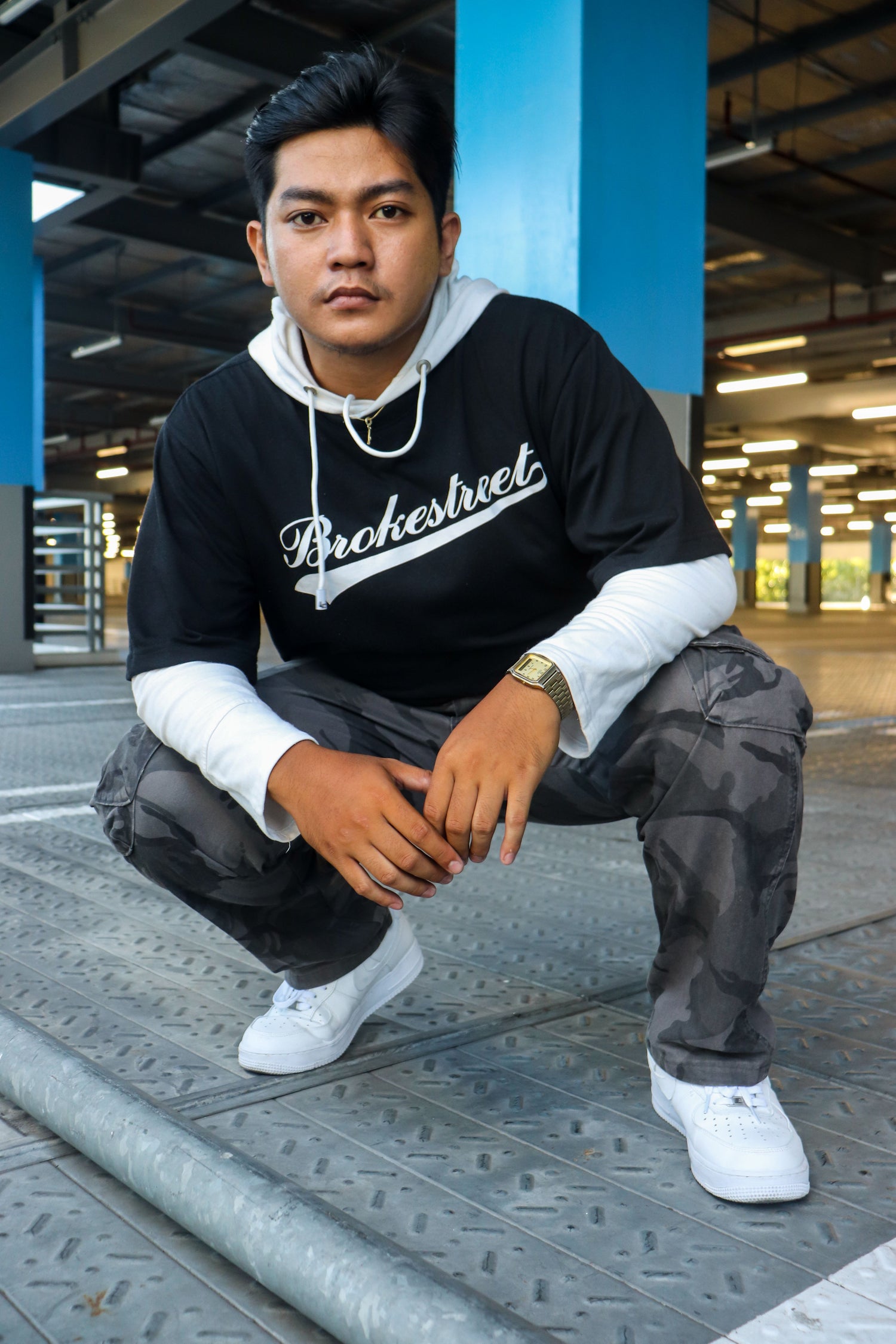 A man crouching in a concrete structure wearing a black and white hoodie with 'Brochestreet' printed across the chest, paired with dark camouflage pants and white sneakers.